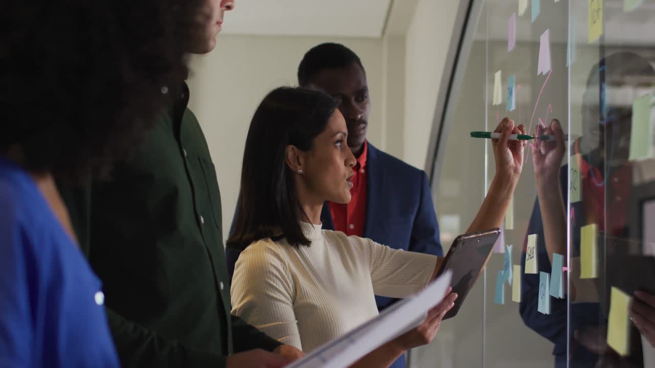 Diverse group of colleagues in meeting room mixed race businesswoman writing notes on glass wall