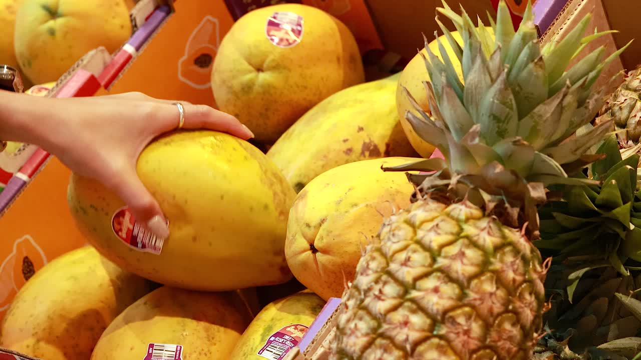 A person examines papayas in a vibrant supermarket setting with bright lighting and colorful produce displays