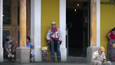 guatemalan indigenous man with traditional clothes stands in a porch outside a local coffee shop along with other locals, as traffic passes by in the street - handheld slow motion