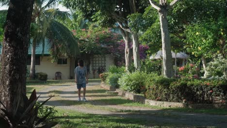 Pretty-young-Asian-girl-cheerfully-walking-and-laughing-on-a-dirt-road-surrounded-by-a-lush-garden-and-trees-during-daytime--wide-static-shot