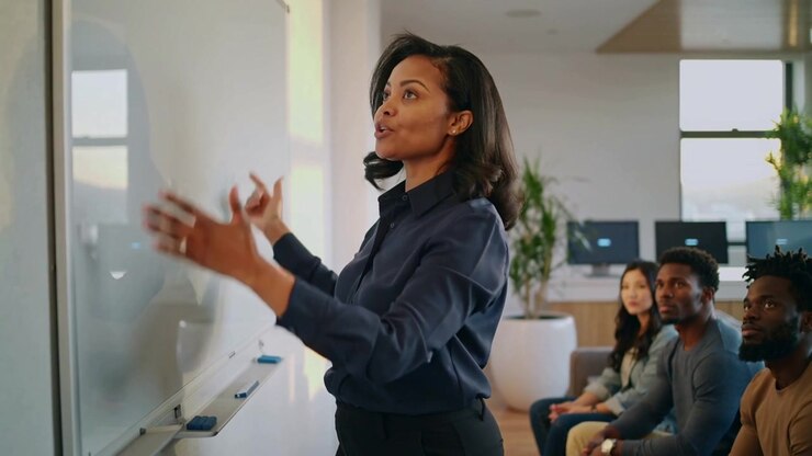 Standing up woman in navy top gesturing, explaining concepts in office with whiteboard, copy space