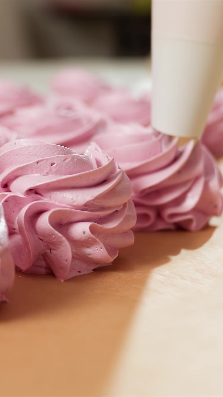 Close-up glasir merah muda yang dibentuk spiral di permukaan. Tekstur mengkilap dan aplikasi yang halus. Berlatar dapur dalam ruangan dengan latar belakang buram yang menonjolkan meringue.