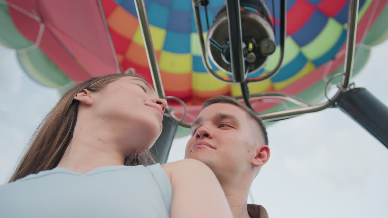Close up of couple standing close inside hot air balloon wicker basket beneath vibrant patchwork envelope panels and braided rope, both gazing upward at metal burner