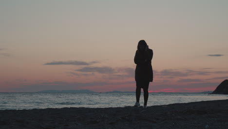 a girl wearing autumn jacket walking along the beach on the sunset with waves of the ocean washing ashore