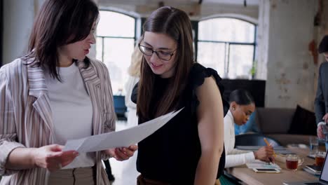 hermosa jefa de negocios caucásica de mediana edad ayudando a una compañera de trabajo más joven con papeles en una oficina de trabajo en el loft de moda