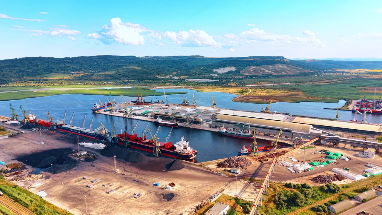 Terminal portuale merci con navi ormeggiate e gru portuali. Vista dall'alto di un porto industriale, cumuli di rottami metallici, acqua blu, colline verdi, cielo sereno.