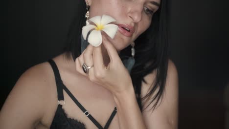 portrait of a sexy brunette woman holding white flower and gently blowing on the petals over black background close up