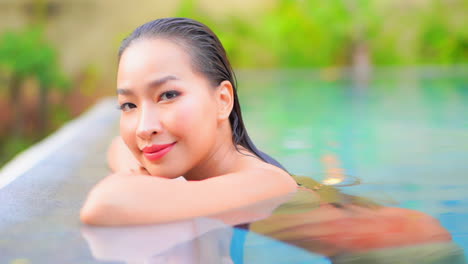 face close-up of asian woman with perfect skin leaning on arms and smiling on border of swimming pool at topical resort in bali daytime