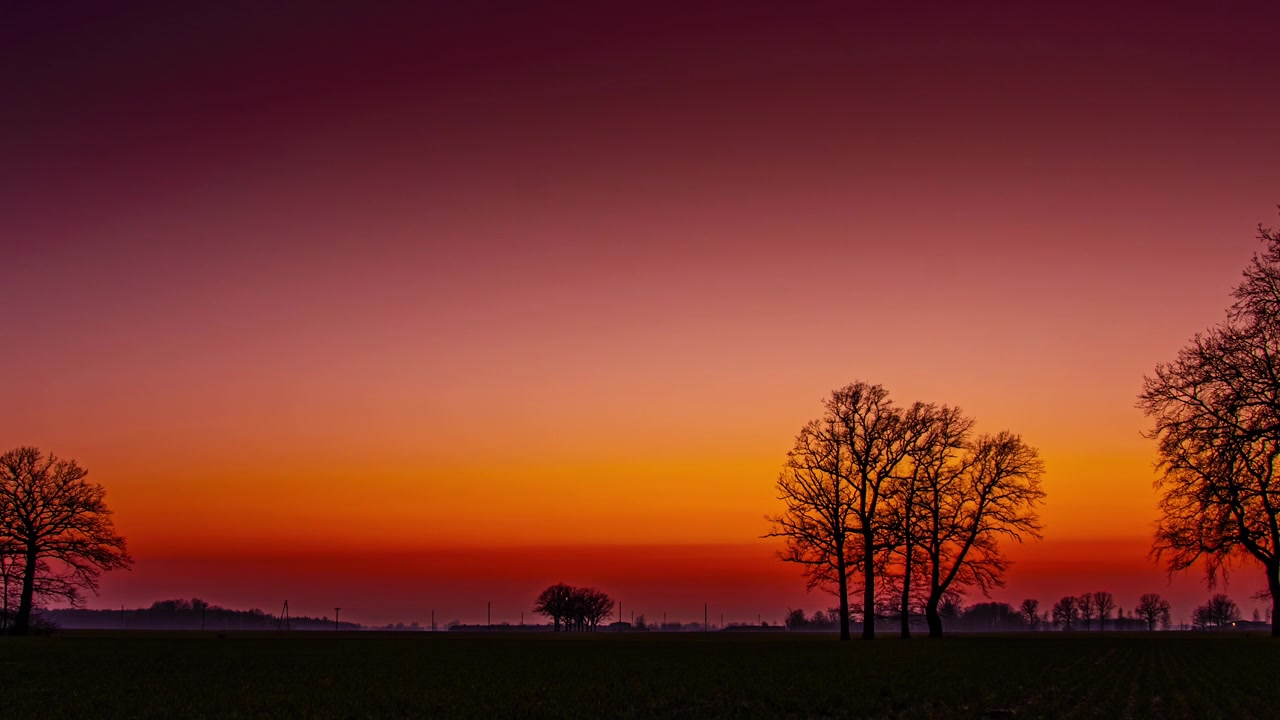 A serene rural horizon where the sky transitions through deep purple and vibrant orange tones.