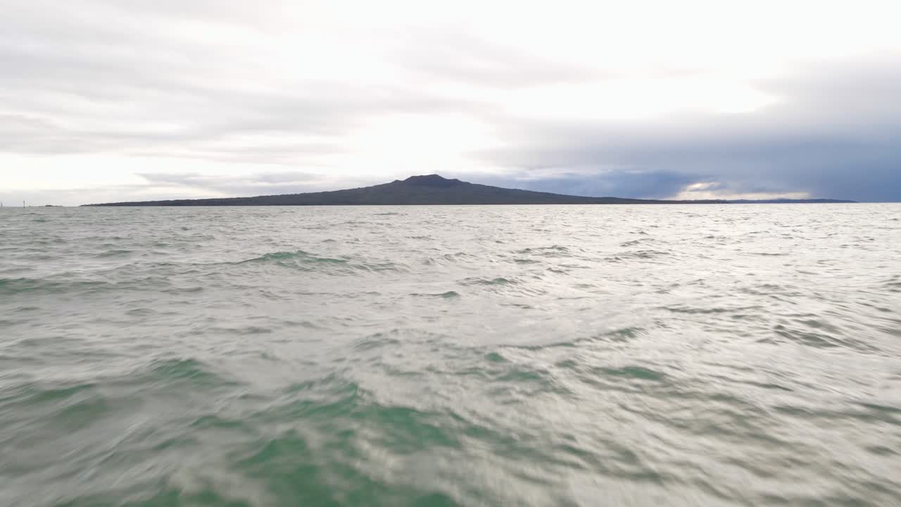 acercamiento aéreo rápido y bajo desde una playa hacia la isla rangitoto durante un amanecer nublado