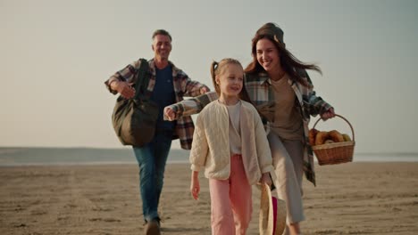 una pequeña chica rubia feliz en una chaqueta blanca corre a lo largo del camino y sus padres la están alcanzando por detrás. y el hombre moreno con cabello gris, la chica morena en una camisa verde a cuadros, durante su caminata y picnic fuera de la ciudad