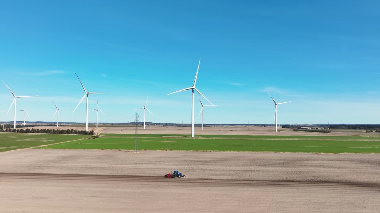 A drone view captures a farmer's tractor working next to a large windmill farm in springtime, with the vibrant fields and towering turbines creating a striking contrast.