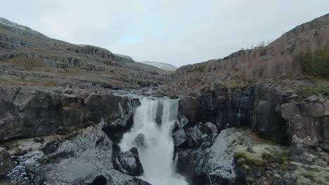 Drone-flying-over-a-beautiful,-wide-waterfall,-towards-a-river-full-of-rocks-in-Iceland