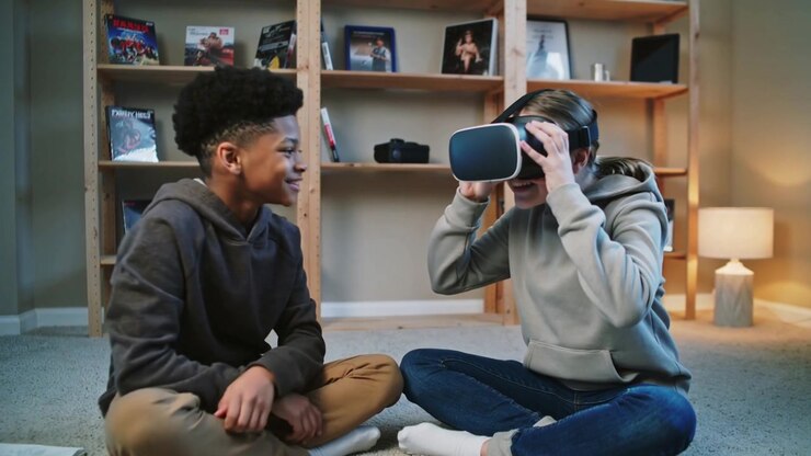 Handing boy passing VR headset to girl at home, both wearing hoodies, helping her explore VR