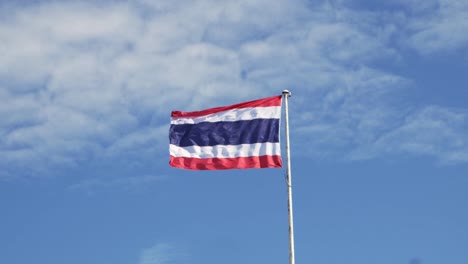 waving the kingdom of thailand flag on a pole with blue sky and white clouds in the background