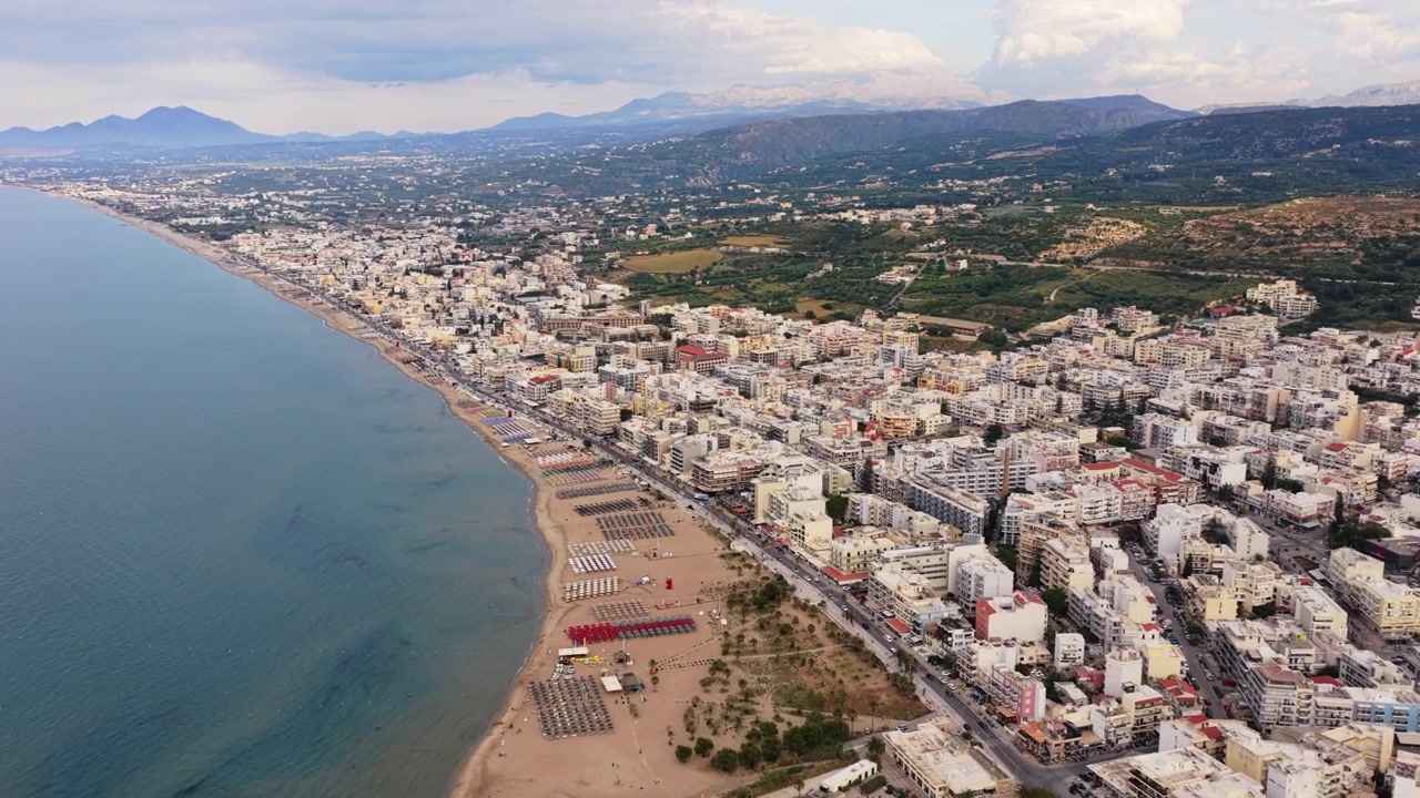 Vista aérea de drone da longa praia de areia da cidade ladeada por guarda-sóis de palha e espreguiçadeiras abaixo da cidade costeira e das colinas, Rethymno, Grécia