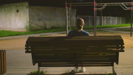 back view of a woman sitting outdoors on a bench at night with light reflecting softly on her face, she holds something on her lap, background features a brick wall under a deep blue night sky