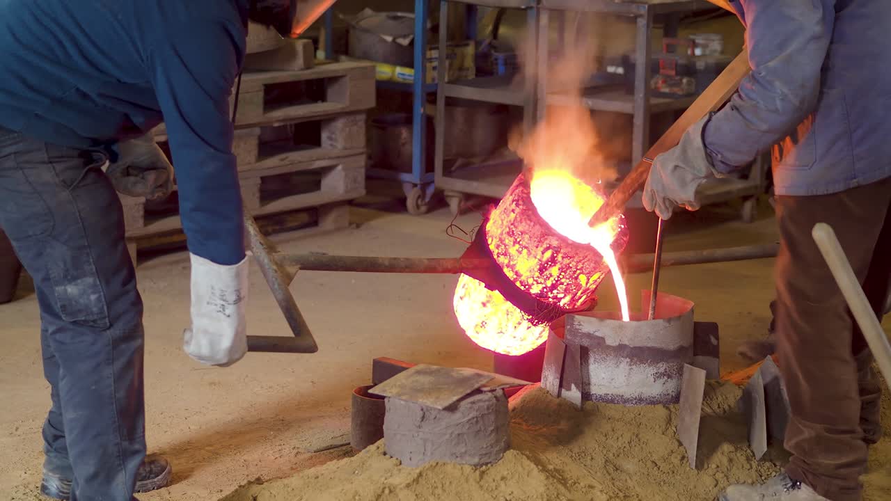 Artisans carefully handle glowing crucible as molten bronze flows into sand mold during traditional bell production using floor casting methods, slow motion