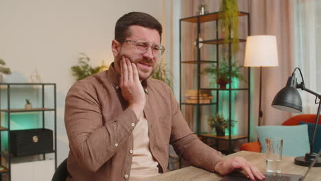 man freelancer sitting at home office drinking cold water feeling toothache touching mouth at home