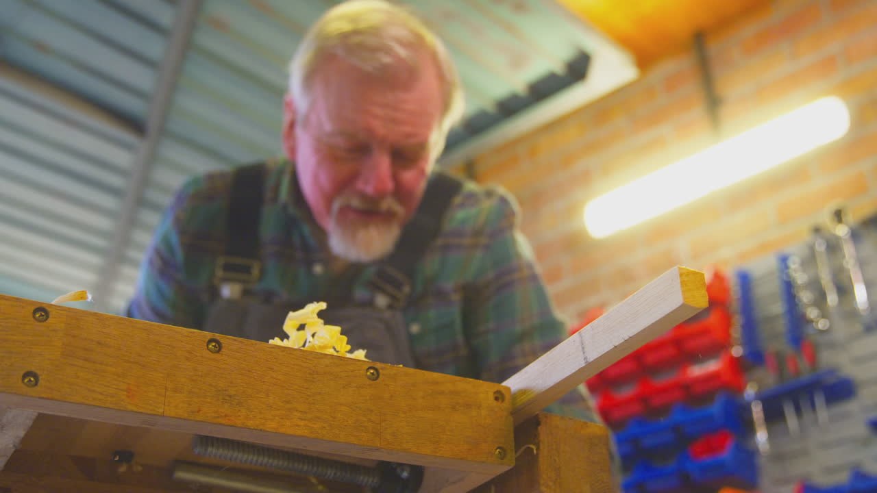 Close Up Of Senior Male Carpenter In Garage Workshop Planing Piece Of Wood