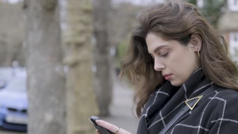 Portrait-of-brunette-businesswoman-standing-on-street-texting-on-her-phone-on-a-cold-cloudy-day-in-central-London