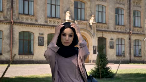 young serious muslim girl in hijab is standing and fix her scarf in daytime in summer, watching at camera, building on background, religious concept