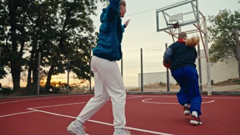 Filmada-Desde-Abajo,-Una-Chica-Rubia-Con-Uniforme-Deportivo-Corre-Y-Supera-A-Todos-Los-Oponentes,-Lanzando-Una-Pelota-De-Baloncesto-Naranja-Al-Aro-En-Una-Cancha-Callejera-Roja-Por-La-Mañana.