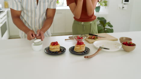 Diverse couple preparing delicious pancakes with strawberries in bright modern kitchen, at home