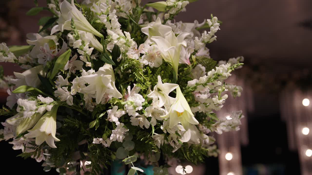 Primo piano scorrevole a destra in slow motion di un elegante centrotavola floreale bianco illuminato a un ricevimento di matrimonio di lusso con sfondo sfocato.