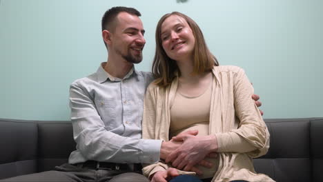 Front-View-Of-Pregnant-Woman-And-Her-Husband-Sitting-On-Sofa-Talking-And-Waiting-For-Gynecologist-In-Medical-Consultation-1