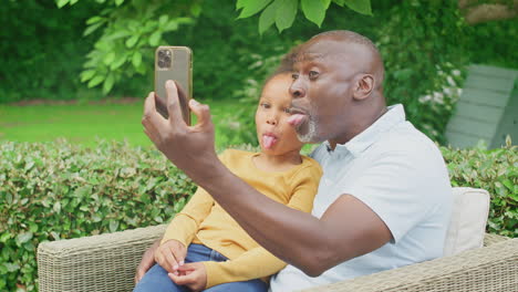 Großvater-Schneidet-Grimassen-Mit-Enkelin,-Die-Im-Heimischen-Garten-Ein-Selfie-Mit-Dem-Handy-Macht