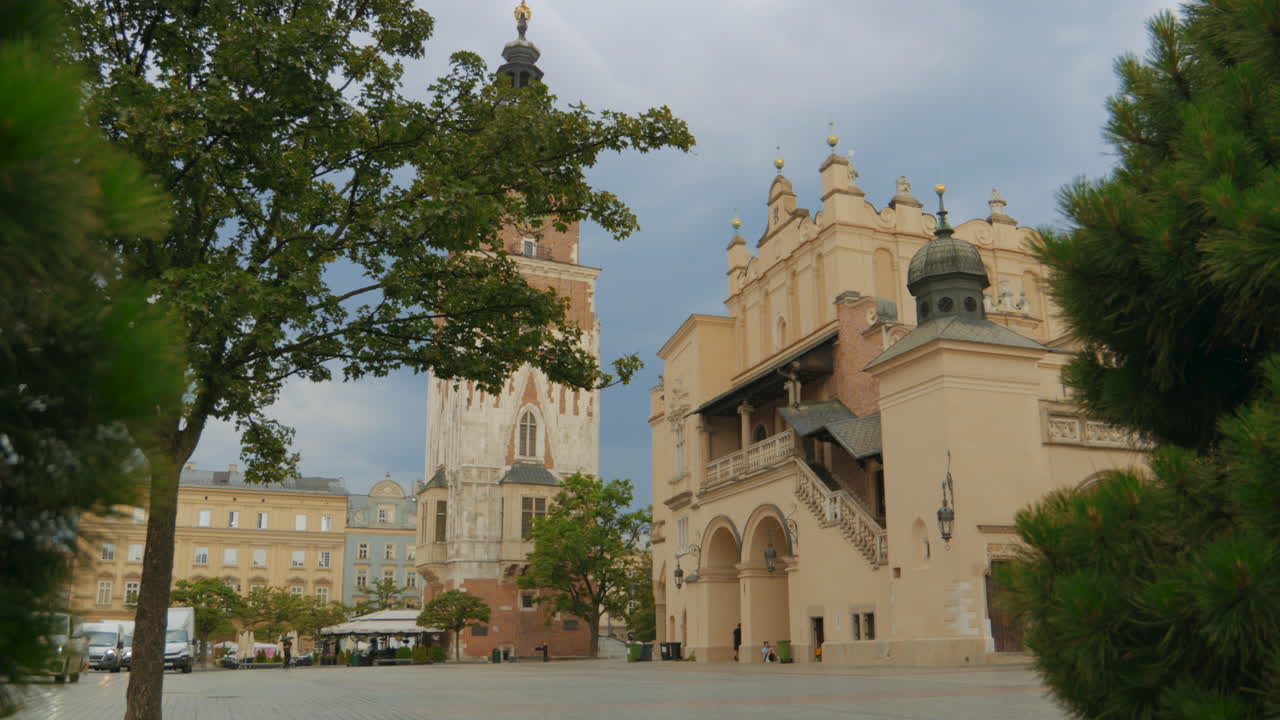 Historic european town square with iconic clock tower and ornate building framed by lush trees on a cloudy day. Cracow Main Square, Poland. Dolly left.
