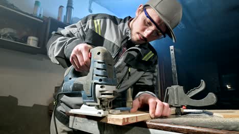Young man with a beard in gray overalls and goggles by profession a carpenter works as an electric jigsaw in his home workshop