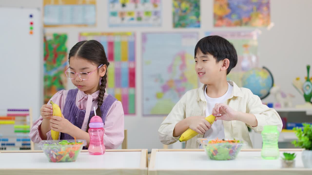 밝은 조명 아래에서 두 명의 아시아 초등학생이 교실 책상에서 바나나를 까서 건강한 간식을 먹고 있어요.