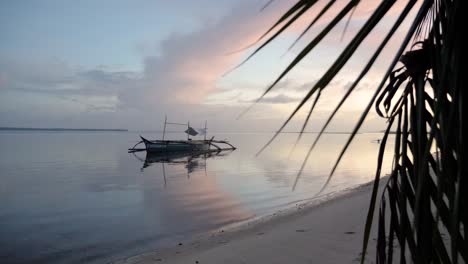 Slow-motion-shot-of-traditional-filipino-bangka-boat-on-calm-waters-at-dusk-as-seen-from-beach-behind-palm-tree