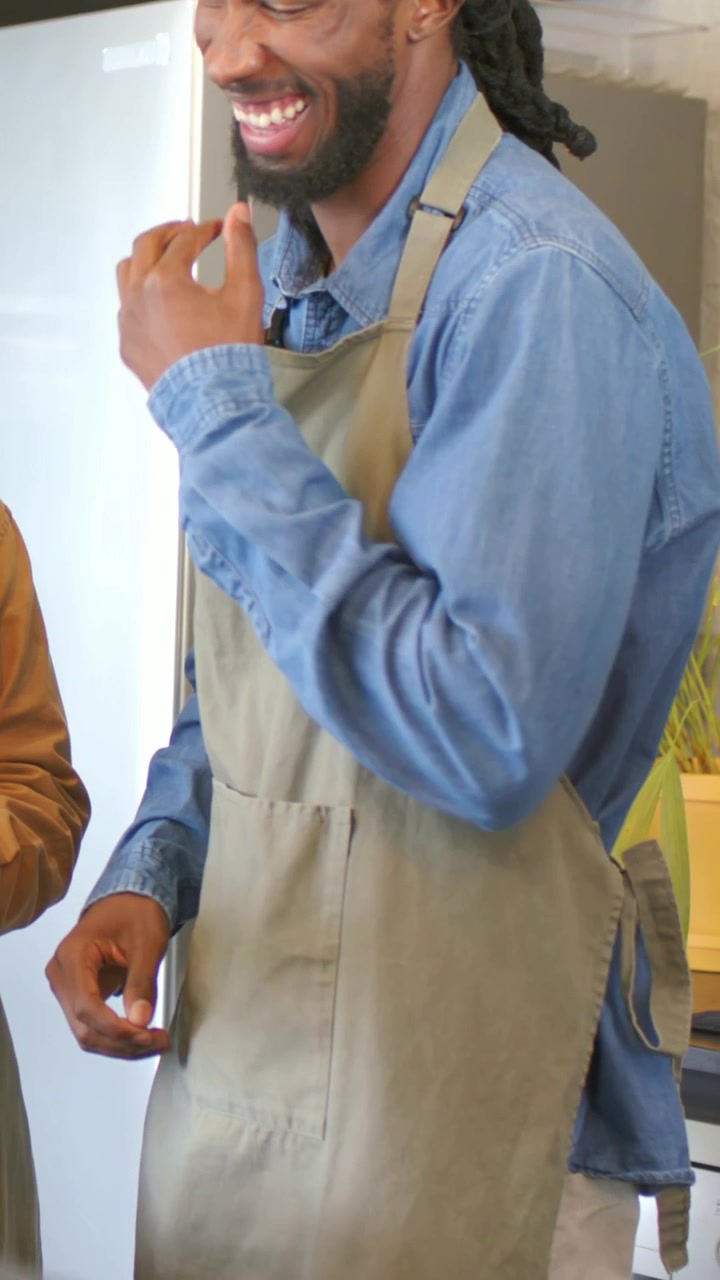 Video verticale: Uomo del caffè che ride, con grembiule di jeans grigio, si tocca il viso, si protende verso il bancone, un collega lo esorta ad aiutare.