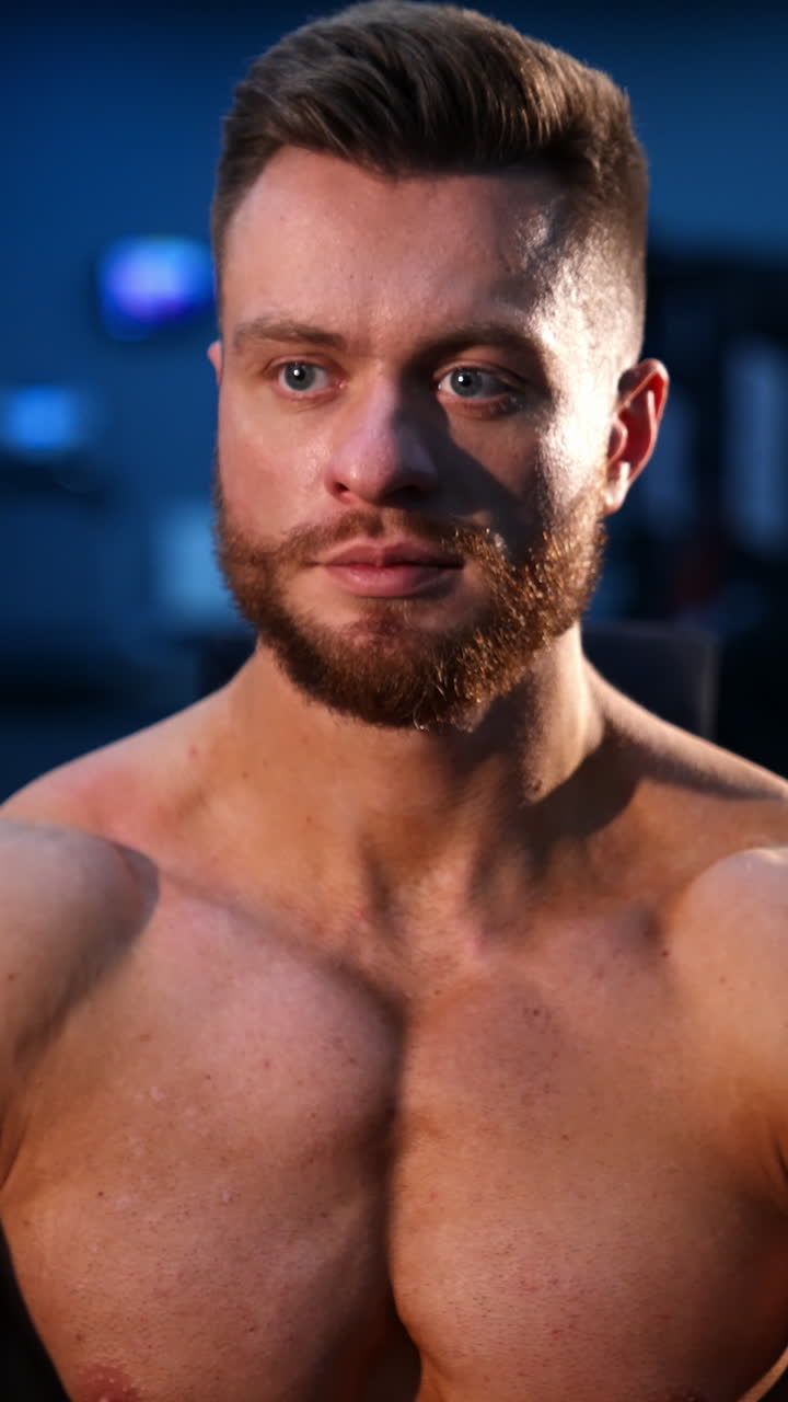 Portrait of bodybuilder indoors. Young bearded sportsman with athletic body sitting in gym. Muscular athlete without shirt. Front view. Vertical video