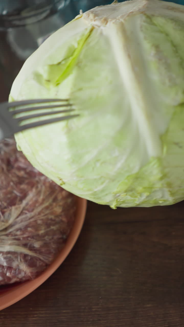 POV footage of a person poking a cabbage head with a spoon and fork in the kitchen, with meat and onion nearby, shot hand-held