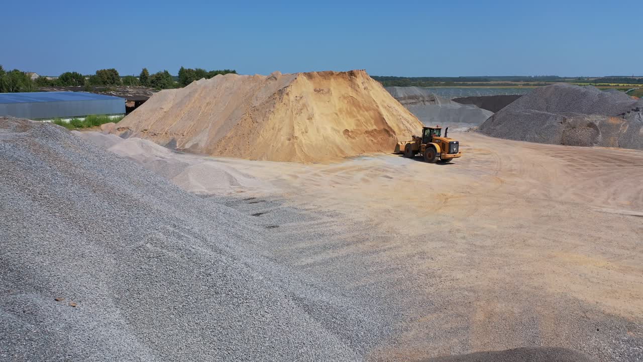 Huge piles of crushed stones and sand outdoors. Bulldozer working on the territory of asphalt plant in a sunny day. Aerial view.