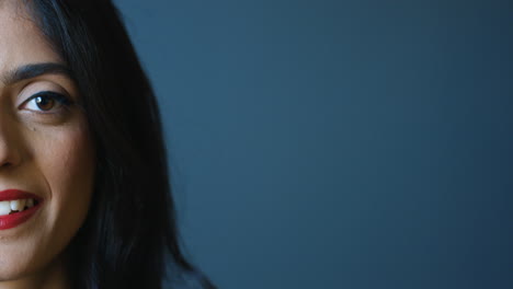 close-up view of half face of happy indian young woman with red lips smiling joyfully at camera on gray wall background