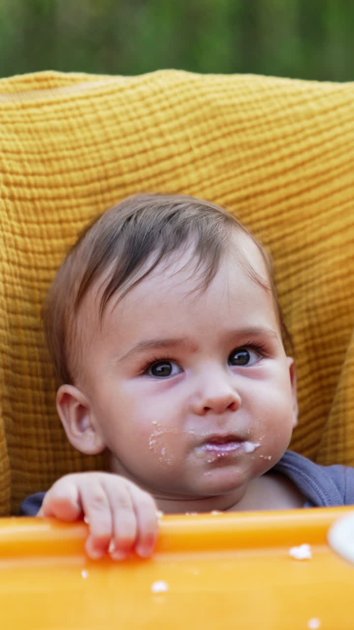 Cute little baby boy in a yellow chair being fed. Mom fills the spoon in plate and gives it to her child. Blurred backdrop. Vertical video