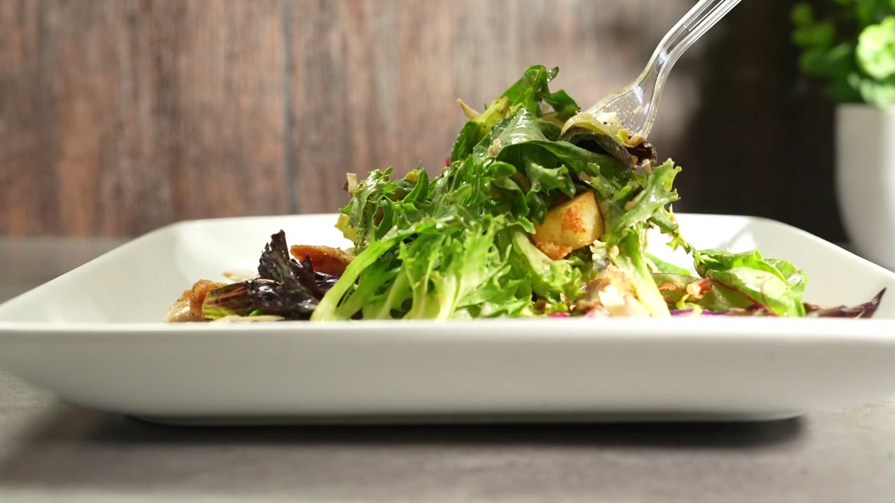 Gennemsigtig plastikgaffel blander en frisk grøn salat med kylling og croutoner, serveret på en hvid firkantet tallerken mod en rustik træbaggrund, der repræsenterer et sundt og lækkert måltid.