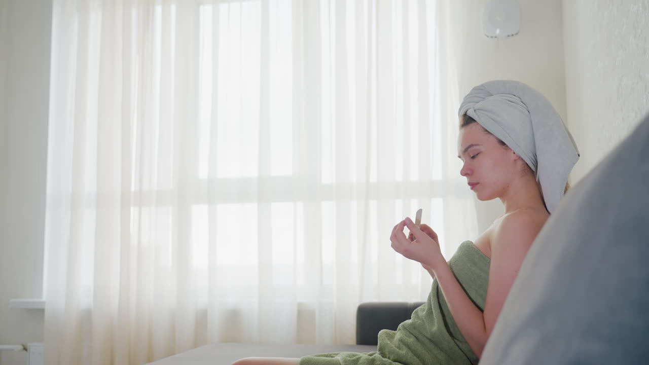 Rear view of woman wrapped in green towel with hair covered by towel turning to sit on couch in softly lit room with sheer curtains, evoking peaceful and clean post-shower moment in home setting