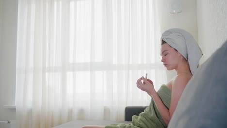 rear view of woman wrapped in green towel with hair covered by towel turning to sit on couch in softly lit room with sheer curtains, evoking peaceful and clean post-shower moment in home setting