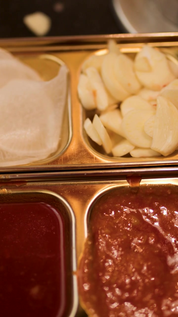 Hand adds fresh garlic slices to metal tray with sauces, pickled radish, kimchi. Warm lighting