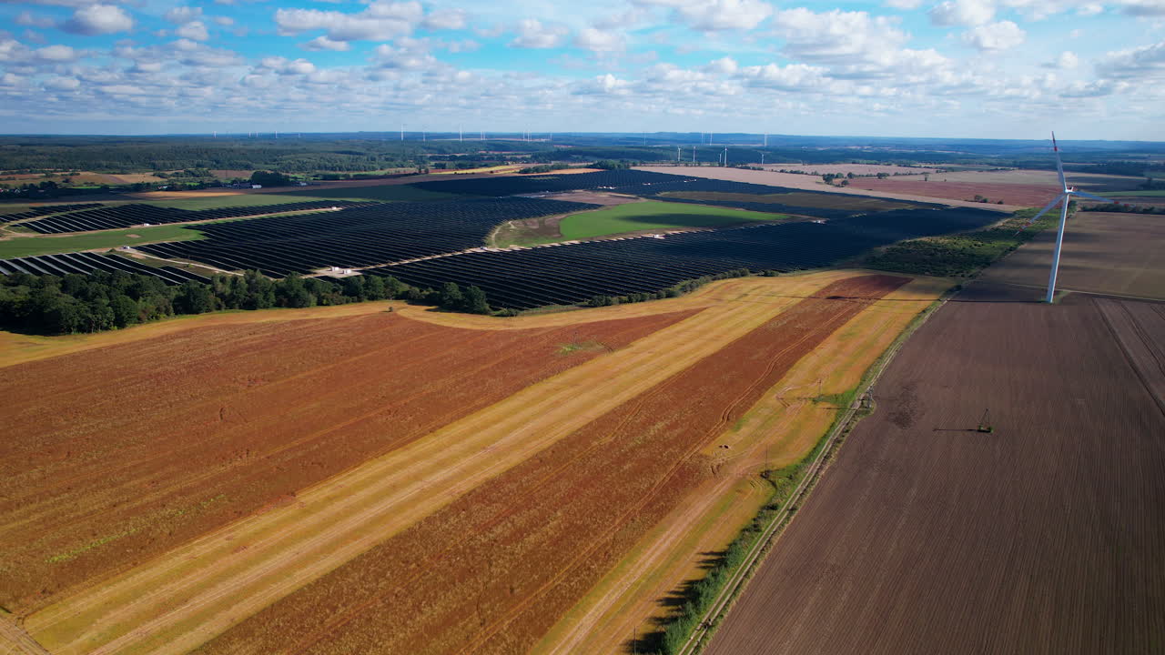 Aerial wide shot showing rural landscape with farm fields, large solar panel units and wind turbines during sunny day