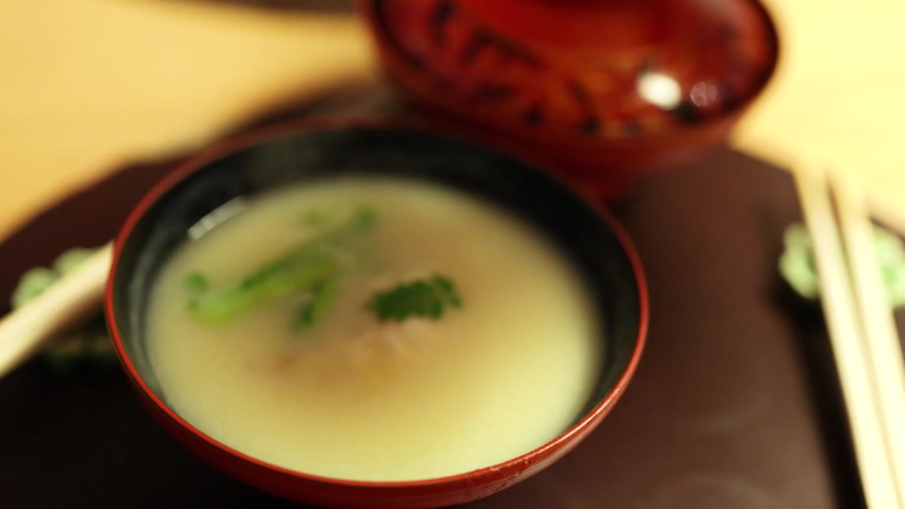 Một bàn tay nhấc nắp của một bát sơn mài đỏ để lộ món súp miso nóng hổi bốc khói