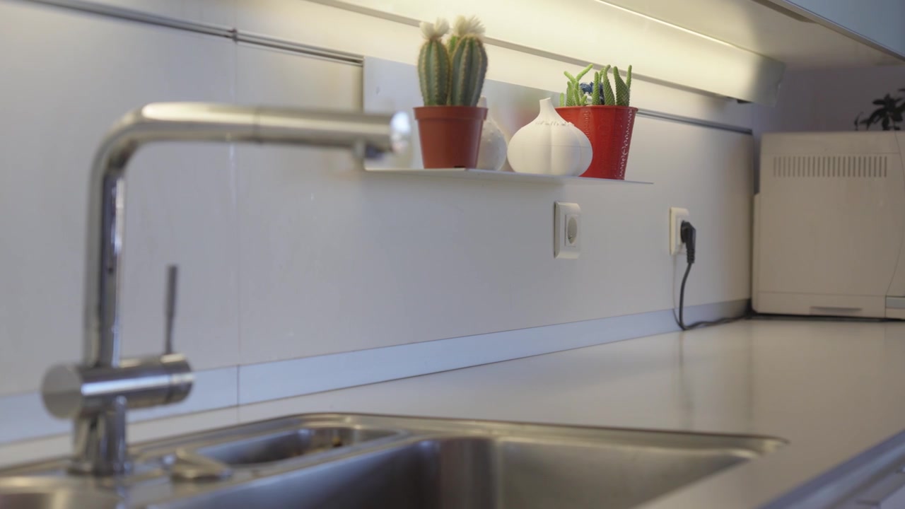 Ripresa ad arco lenta di un moderno lavello da cucina con rubinetto cromato, paraschizzi piastrellato bianco, piccole piante in vaso ed elettrodomestici in un ambiente interno luminoso