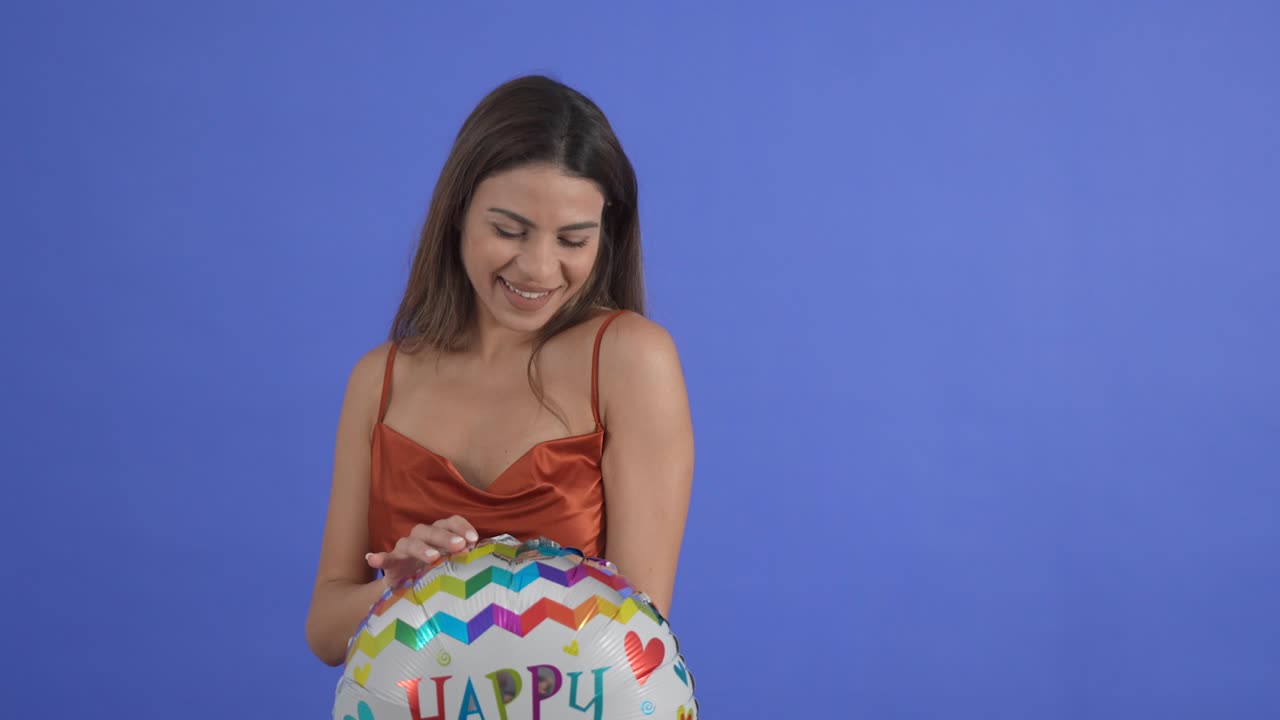 una mujer joven posa con un globo de feliz cumpleaños, aislada en un fondo de estudio azul