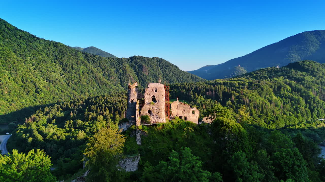 Castle ruins standing on the hill. Mountains covered with beautiful forest are around the sight. Aerial view.